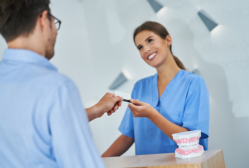 The image shows a woman wearing a blue uniform and smiling at a man who is handing her a card, with a dental implant on display behind them.